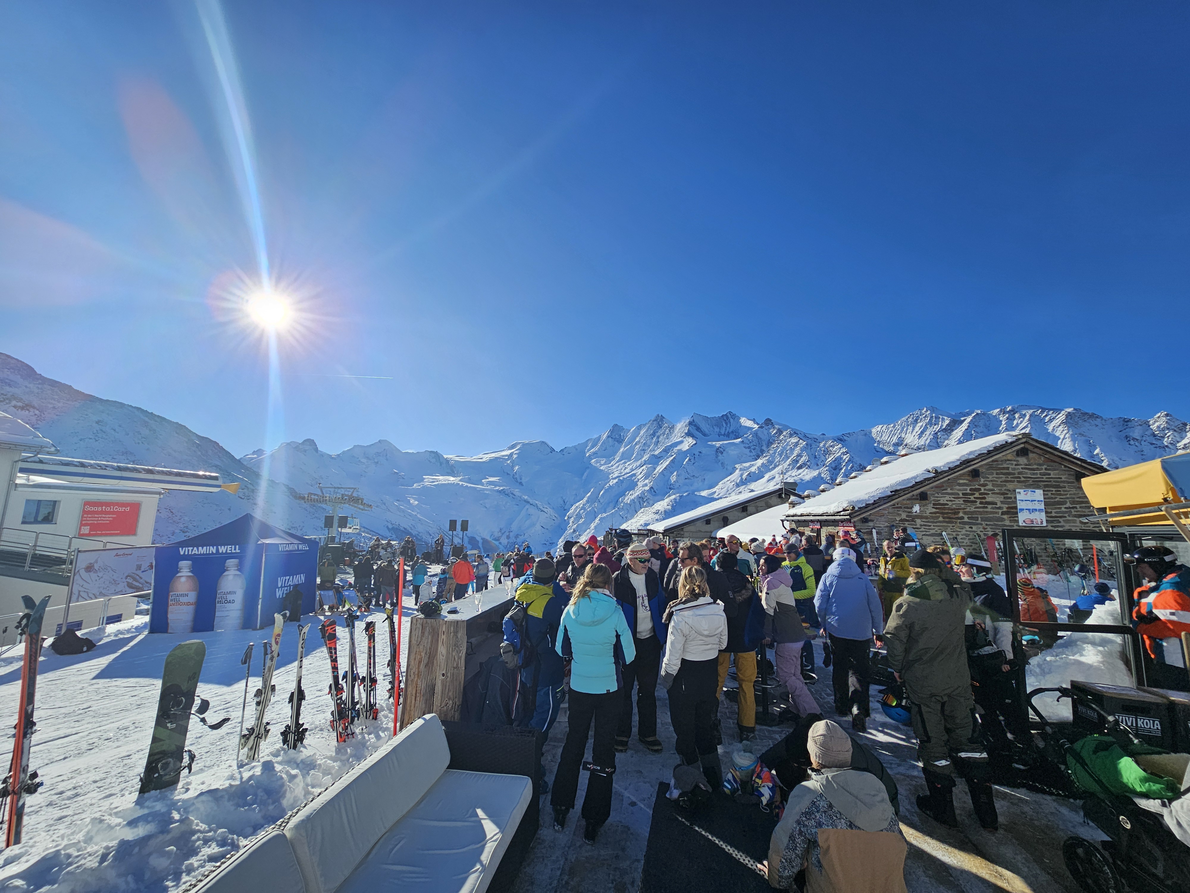 Personen geniessen die Aussicht auf einer sonnigen Bergterrasse in verschneiter Winterlandschaft. Im Hintergrund sind schneebedeckte Berge, blauer Himmel und helles Sonnenlicht zu sehen.